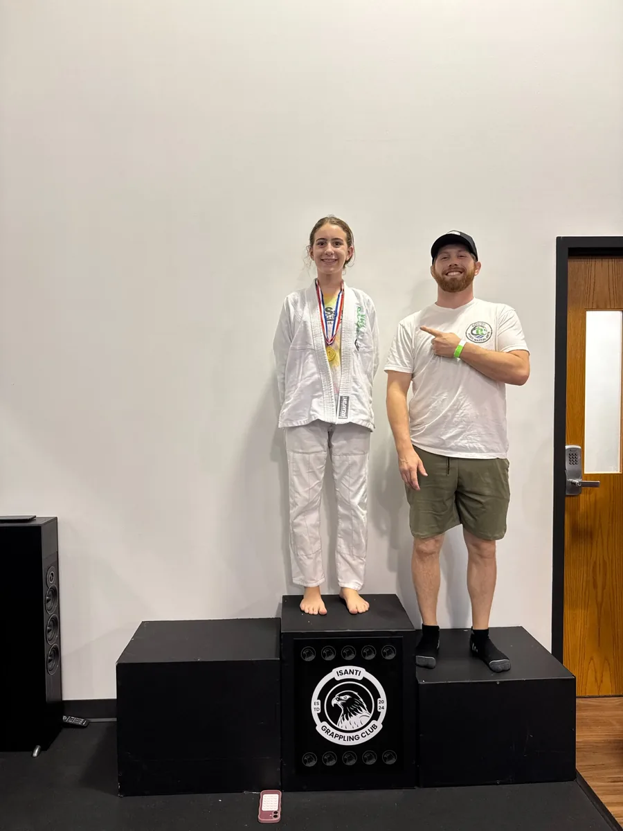 A teen wearing a tournament medal on the podium beside a Roots coach.