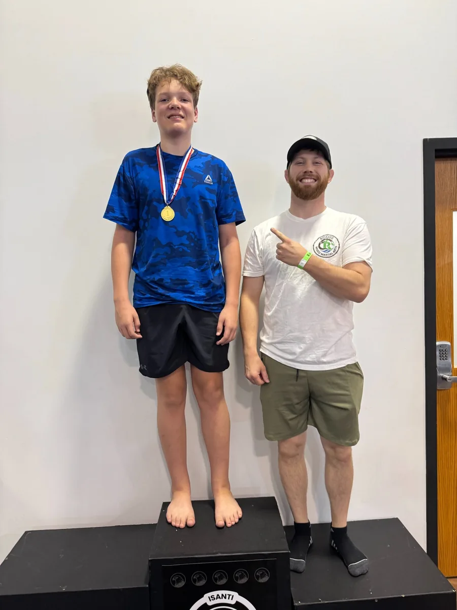 A teen in a blue shirt wearing a tournament medal beside a Roots coach.